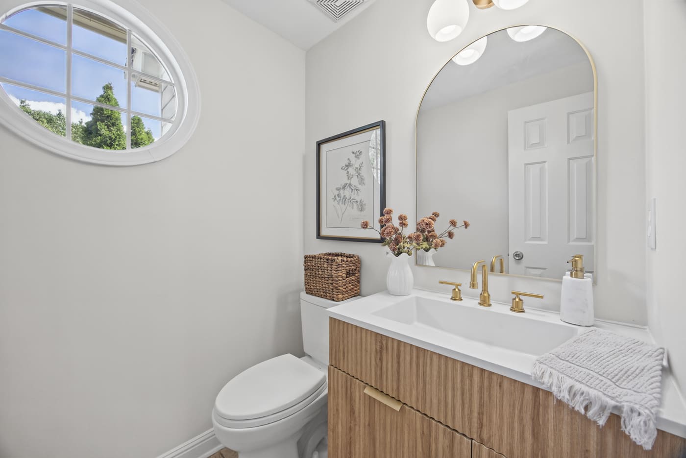 The powder room with a wood-front vanity, brass fixtures, an arched mirror and a round window.